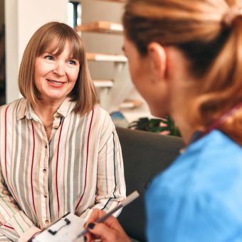 Happy young female nurse provide care medical service help support smiling mature woman at homecare medical visit, lady carer doctor give empathy encourage retired patient sit on sofa at home hospital